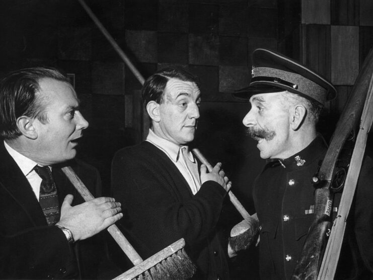 Denholm Elliott and Bryan Pringle holding brooms while talking to Bert Croucher