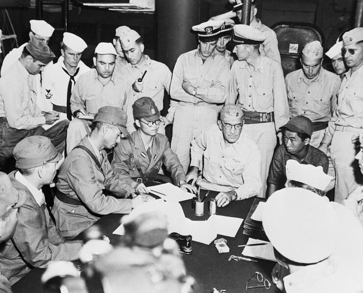 US and Japanese officials standing around Shigematsu Sakaibara, who's sat at a table