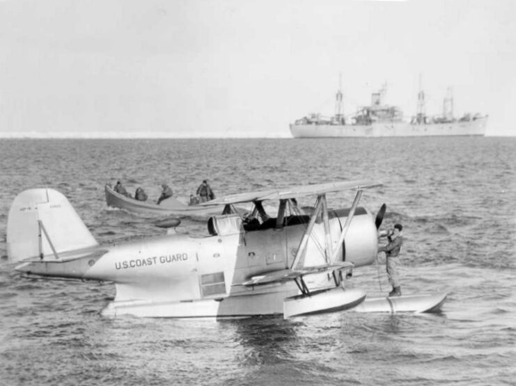 Grumman J2F Duck floating on the ocean's surface, with a ship sailing by in the distance and a smaller watercraft anchored nearby