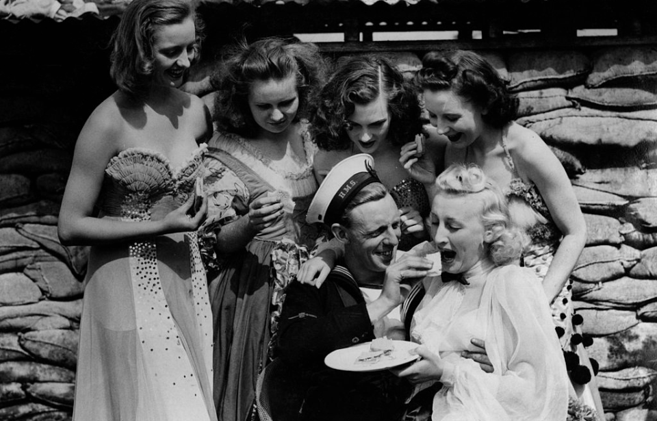Royal Navy sailor feeding cake to a Windmill Girl while four other women watch.