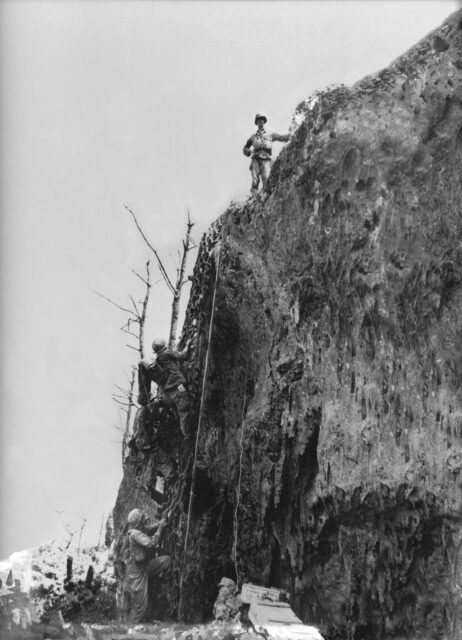 Desmond Doss standing at the top of the Maeda Escarpment as other soldiers climb up to him