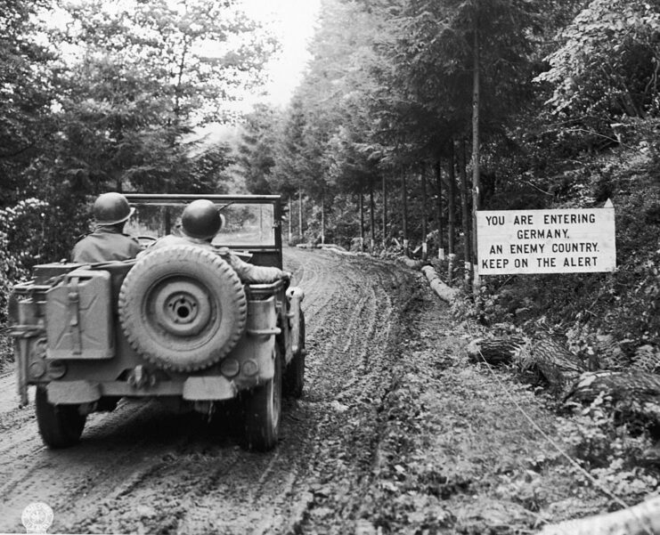 US Army Signal Corps troops driving a Willys MB past a sign, which reads, "YOU ARE ENTERING GERMANY, AN ENEMY COUNTRY, KEEP ON THE ALERT"