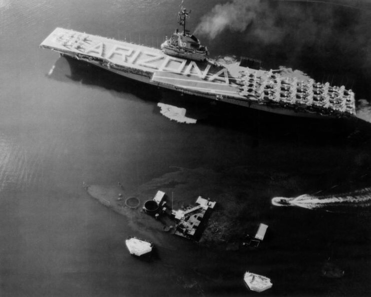 Aerial view of the USS Bennington (CV-20), with "ARIZONA" written on her flight deck, transiting past the wreck of the USS Arizona (BB-39)