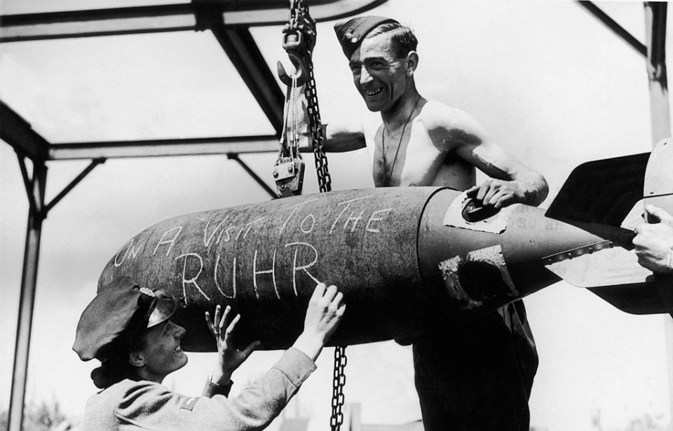 A Royal Air Force (RAF) airman and Women's Auxiliary Air Force (WAAF) member standing with a bomb with "ON A VISIT TO THE RUHR" written on it in chalk