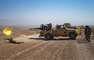 Soldiers standing around in the Syrian desert while another and a military vehicle fire their weapons