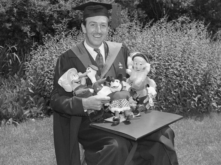 Walt Disney dressed in graduation regalia while holding dolls of his cartoon characters