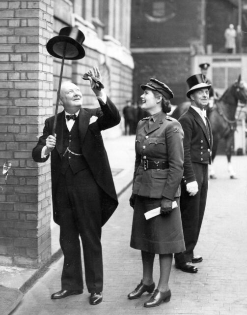 Winston Churchill balancing his top hat on his cane while his daughter, Mary, watches