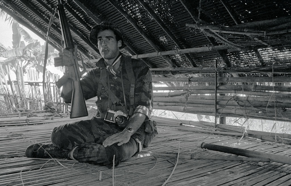 Sean Flynn holding up a weapon and sitting cross-legged in a hut
