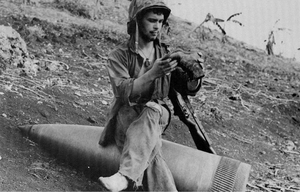 Pfc. Raymond Hubert sitting on a naval shell while emptying sand from his boot