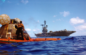 Frogman standing with the Apollo 17 Command Module, with the USS Ticonderoga (CVS-14) in the distance