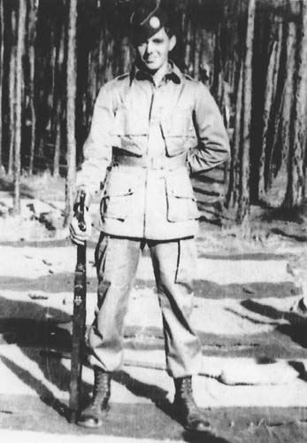 Darrell Powers standing in a wooded area in his uniform and with a rifle