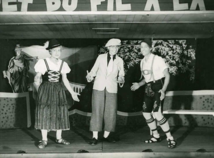 Two men dressed as Hansel and Gretel, with a third standing between them in a hat, shirt and blazer, tie and slacks