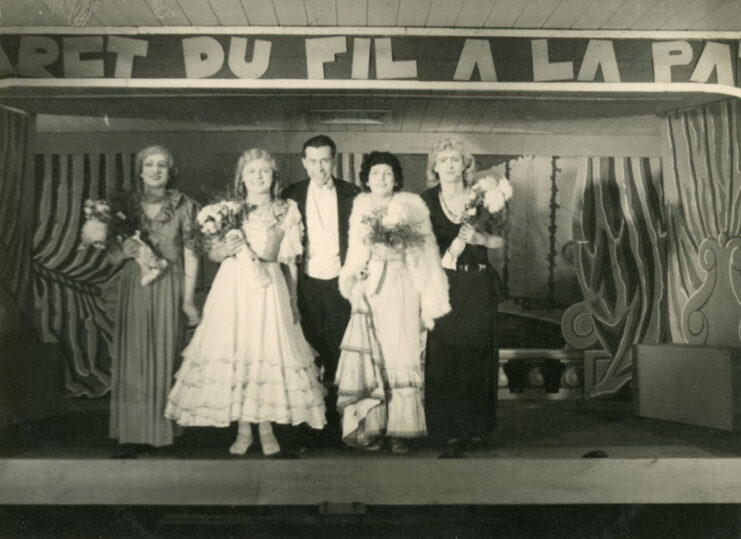 Four servicemen dressed in drag while standing on stage, with a fifth standing between them in a suit