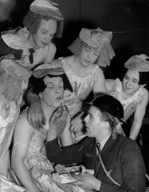 Man applying lipstick to a performer dressed in drag, while five others stand around them