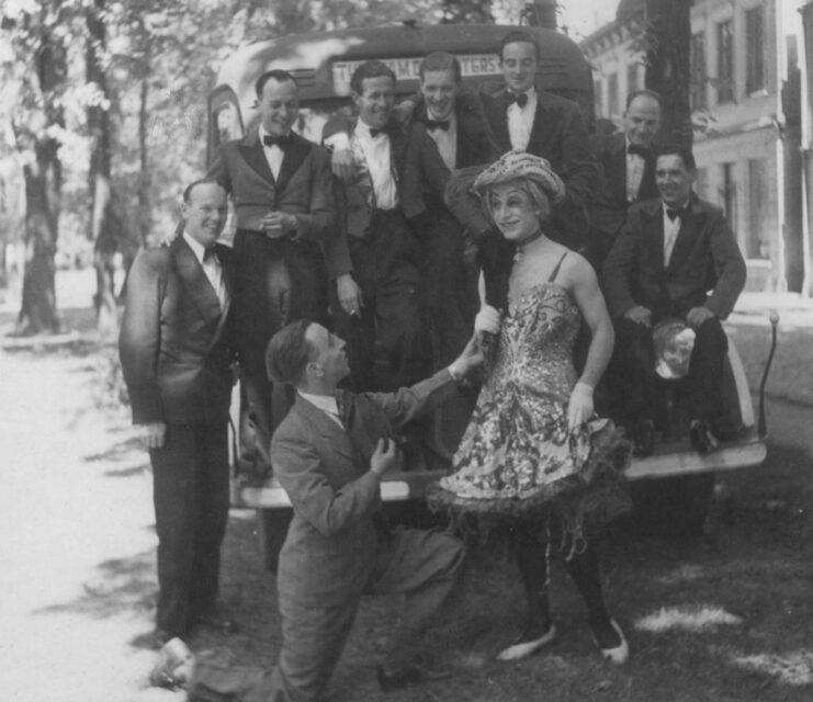 Group of men in suits stand on the back of a truck while another man, dressed as a woman, stands with them.