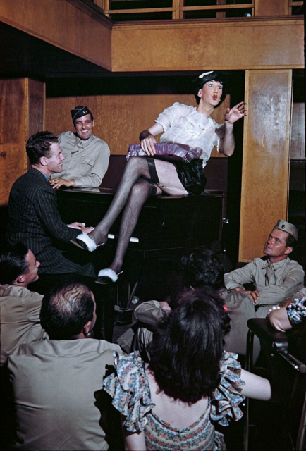 Soldier sitting on a piano dressed in drag while others sit around him