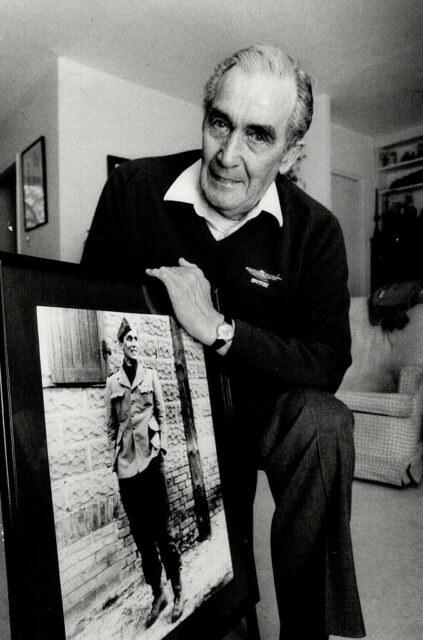 Wally Floody standing with a photo of him in his military uniform during the Second World War
