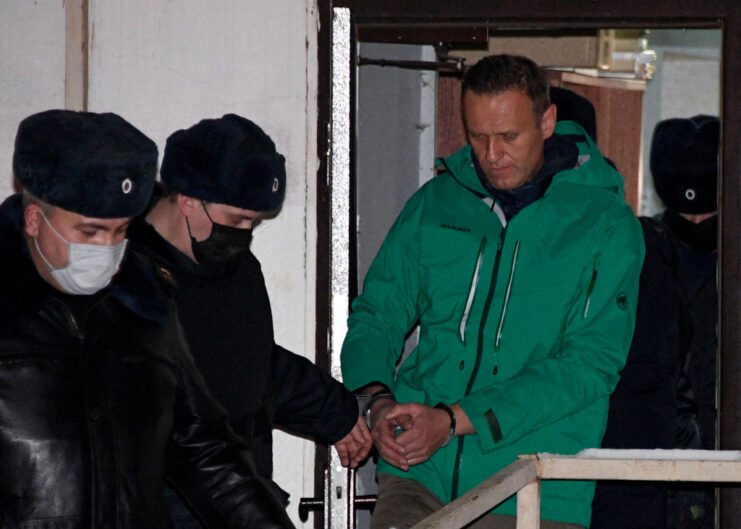 Two Russian police officers leading Alexei Navalny, who is handcuffed, through a door