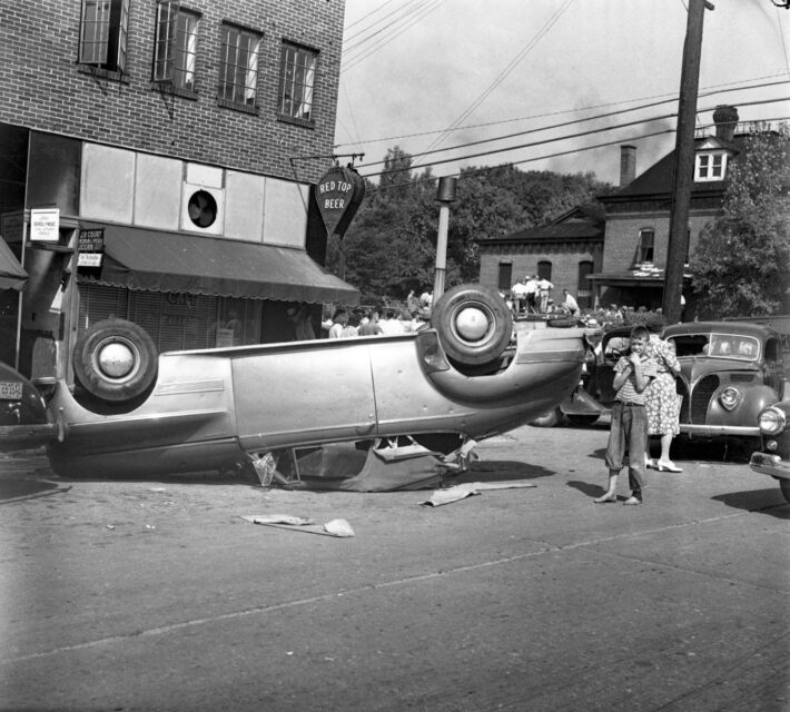 Child and his mother standing beside an overturned car in the middle of a street