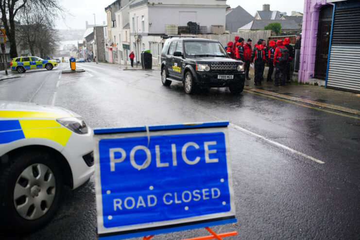 Bomb disposal experts standing near vehicles and a "POLICE ROAD CLOSED" sign