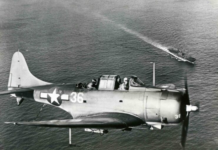 Douglas SBD-3 Dauntless in flight over the ocean, with an aircraft carrier transiting below