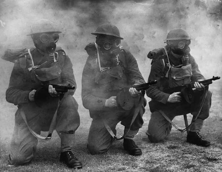 Three British soldiers in gas masks crouching while holding Tommy Guns