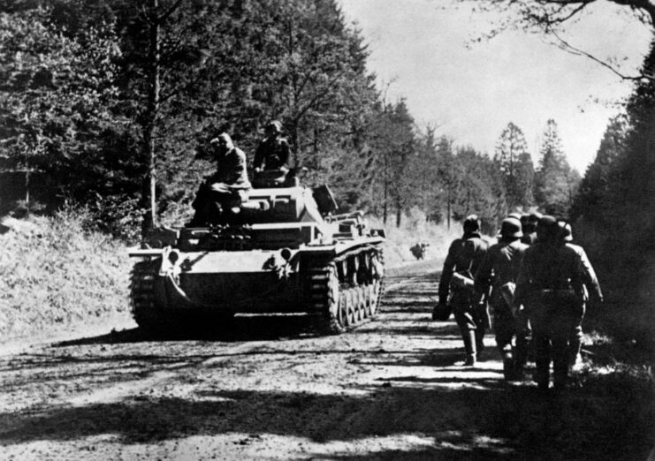 Sappers walking along a dirt road while a tank drives by