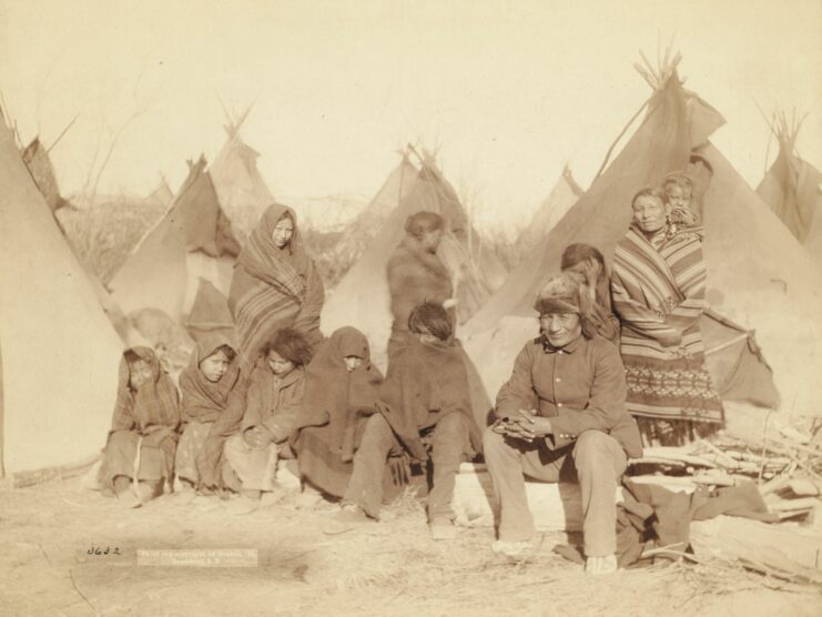 Black and white, 5 children wrapped in blankets and one man sit on a log, three women wrapped in blankets stand behind them. One woman has a child on her back. Tents can be seen in the background.