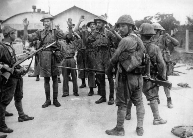 British troops holding their hands up while Japanese soldiers aim their rifles, equipped with bayonets, at them