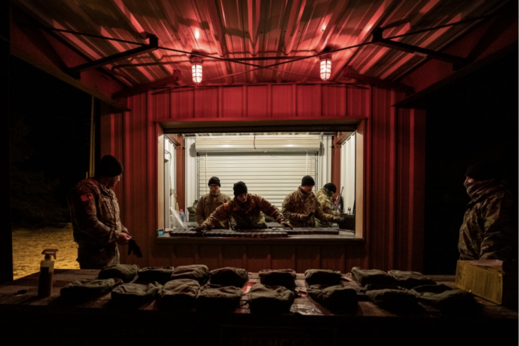 Dark room with two red lights on the ceiling. Ammo and gear on the table in the middle of the room. two men in uniform stand on either side of the table and four men stand in a room with the lights on in the middle of the picture and look through a window into the dark room.