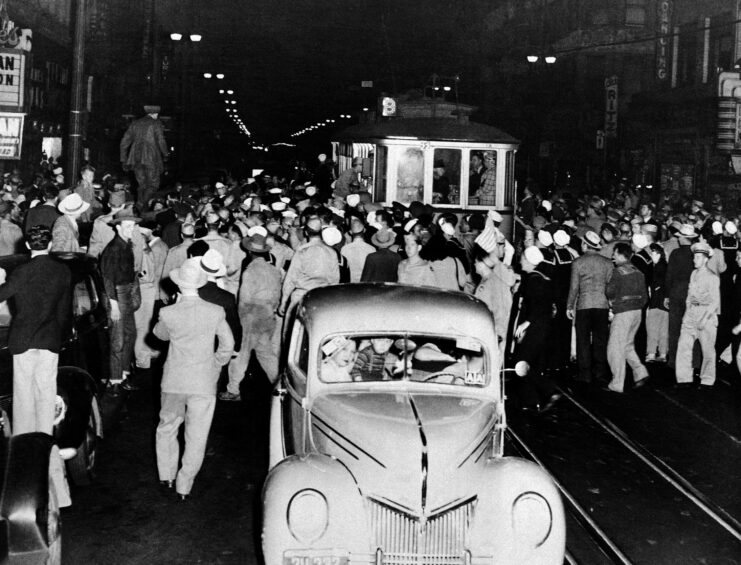 Black and white, car in centre of image with streetcar and streets filled with people behind. Two people look out of windshield of car. In dark street. 