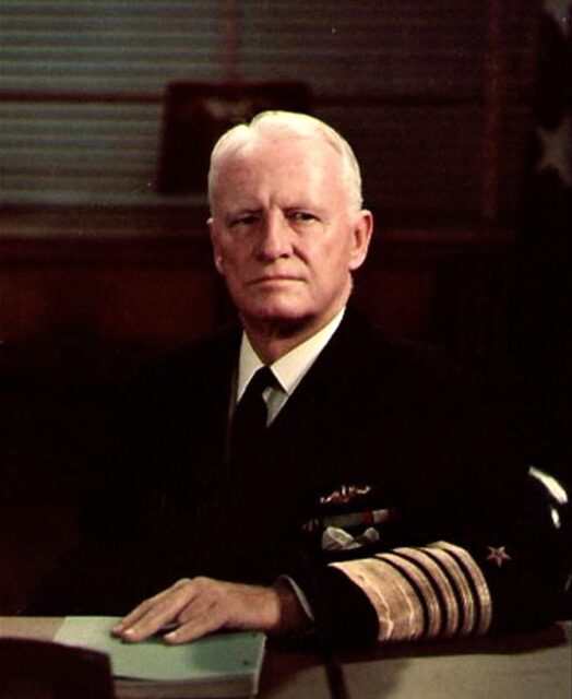 Man with white hair sitting behind a desk while wearing a naval uniform. One hand resting on the desk.