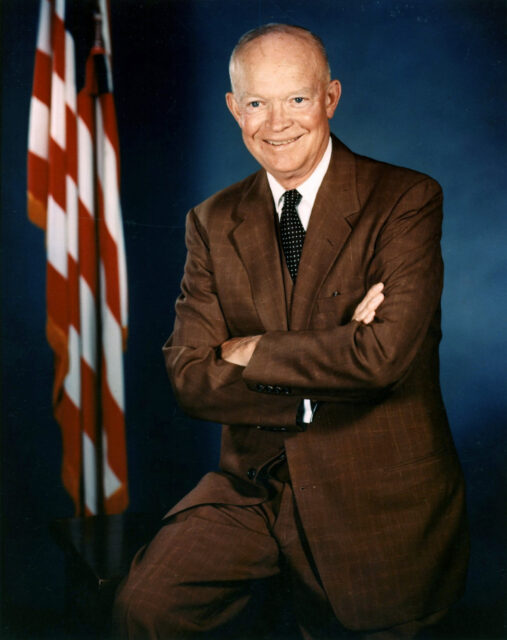 Man in brown suit stands in front of a blue background and an american flag, smiling with arms crossed.