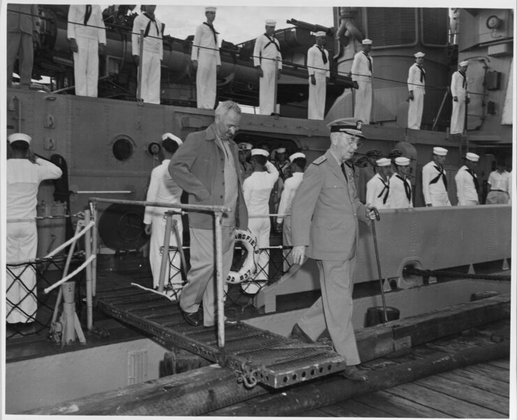 One man in a naval uniform and one man in a suit step off of a boat onto a dock as sailors on deck in sailor uniforms stand at attention and salute on the boat behind them. Black and white.