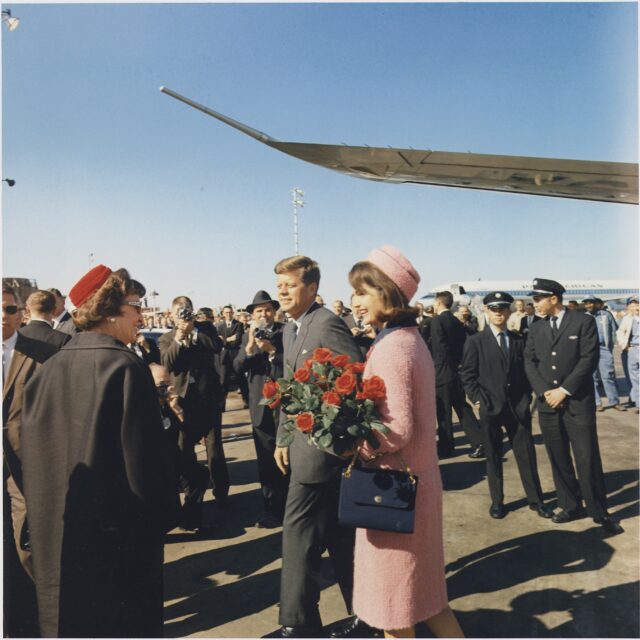 Brunette woman holding red roses and wearing a pink skirt suit and hat, smiling beside man wearing a suit and talking to a woman in a black coat and red had. WIng of a plane in the background and men in suits and uniform crowd behind the figures.