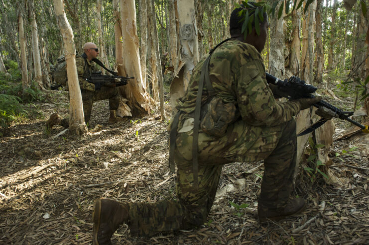 Two US Army soldiers in camo kneeling in the jungle holding guns.