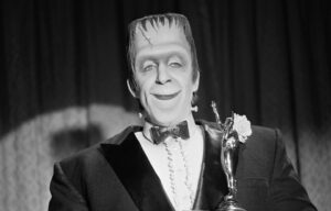 Fred Gwynne as 'Herman Munster' stands in a tuxedo before a curtain with an award trophy in a still from the CBS television situation comedy 'The Munsters' episode 'Will Success Spoil Herman Munster?.'