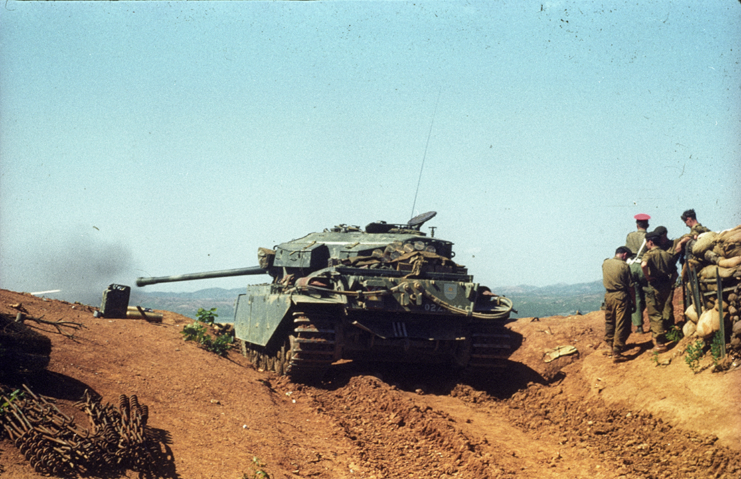 Soldiers standing near a Centurion tank, which is parked on a dirt road