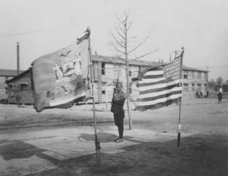 Soldier standing between the American flag and the flag of the former 15th New York National Guard Regiment