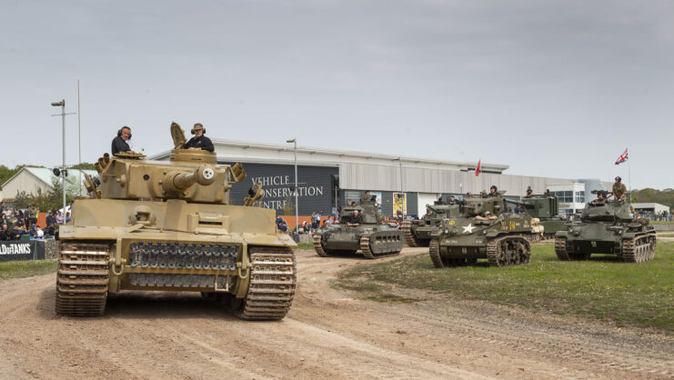 Various military tanks driving along a dirt track and parked in a grassy area