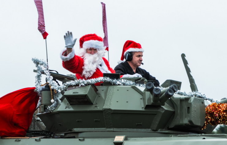 Man dressed as Santa Claus sitting atop a tank, with a man next to him wearing a Christmas hat