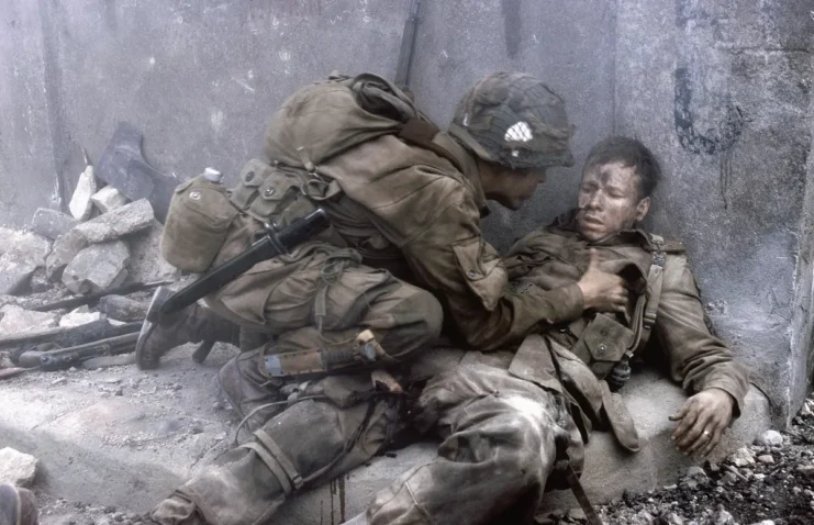 A movie still from the Band of Brothers (2001) movie of a soldier helping another soldier who has fallen on his back against a wall.