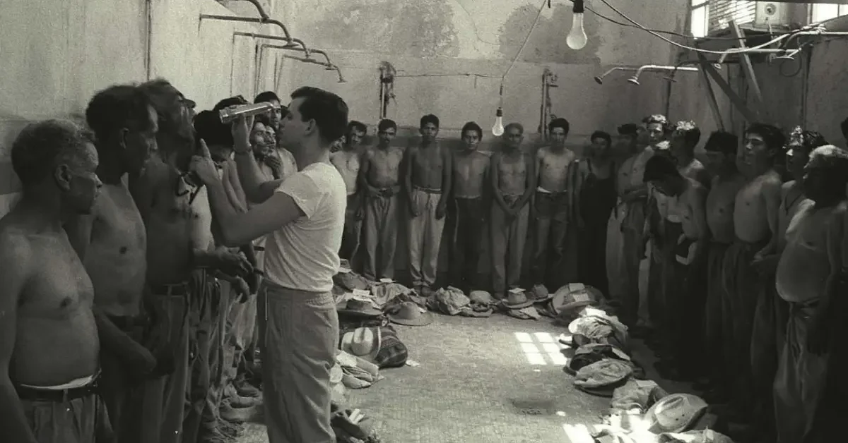 An official examines a bracero's teeth and mouth with a flashlight while others stand next to him with their backs to the wall at the Monterrey Processing Center, Mexico, 1956